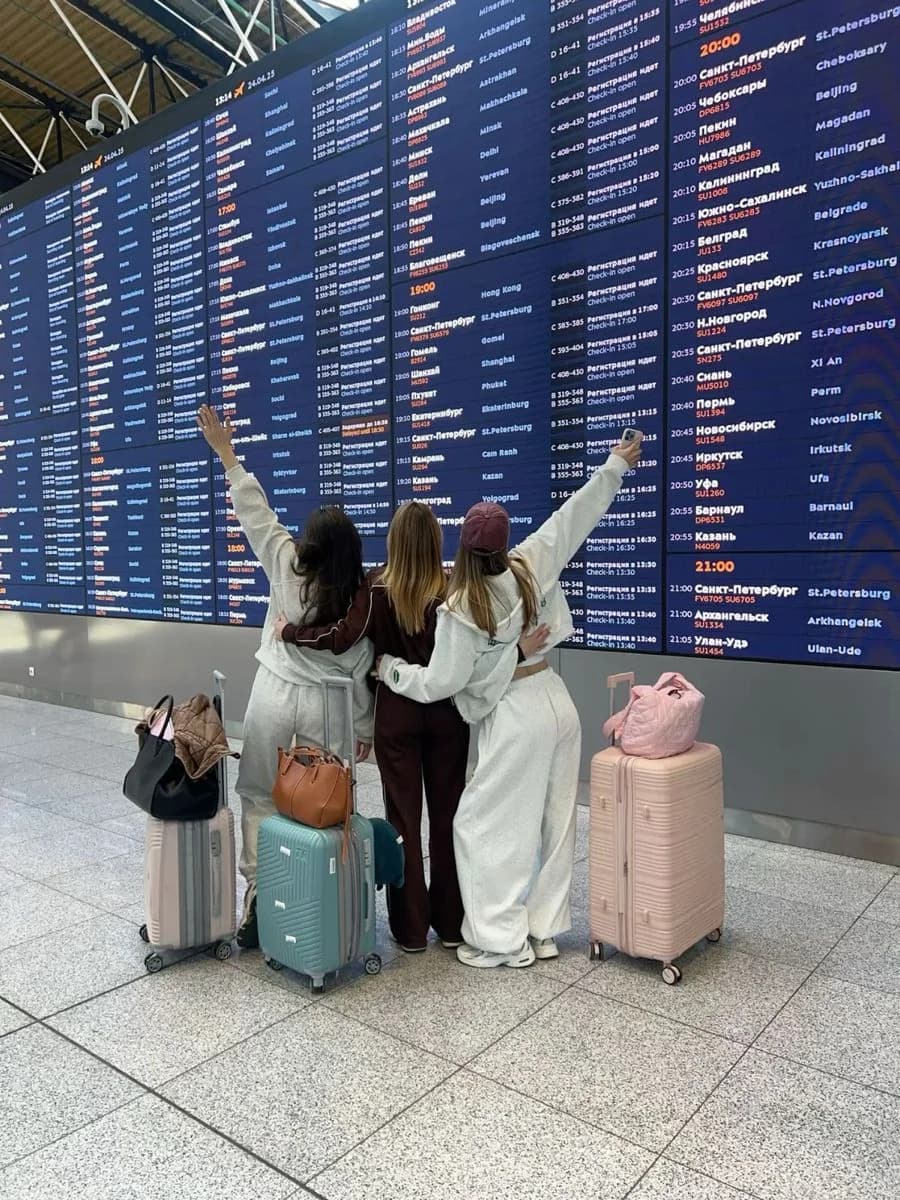 Three women with luggage waiting beneath an airport departures board
